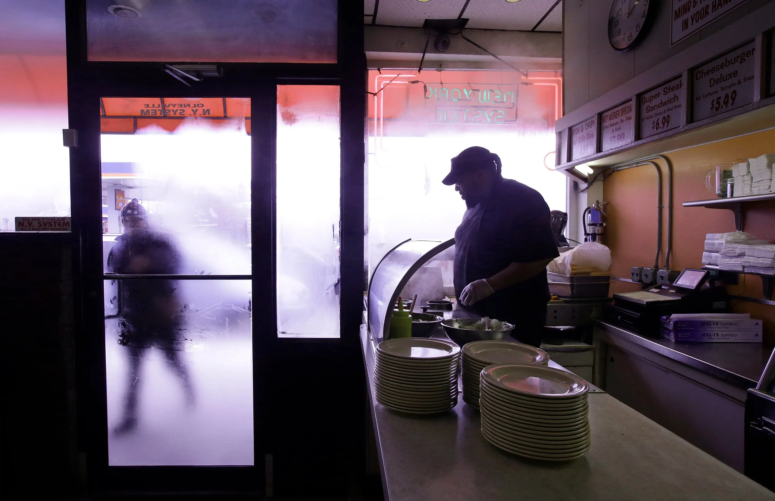Cold Day… Hot Wieners — In this Monday, March 3, 2014 photo, Nicholas Barros, a waiter and cook at “Olneyville New York System of Providence” restaurant works the grill as a customer walks up to the door in Providence, Rhode Island. The James Beard Foundation named the Rhode Island restaurant one of five 'American Classics' that year as best known for hot wieners, also called hot dogs.  (AP Photo/Stephan Savoia)  © The Associated Press