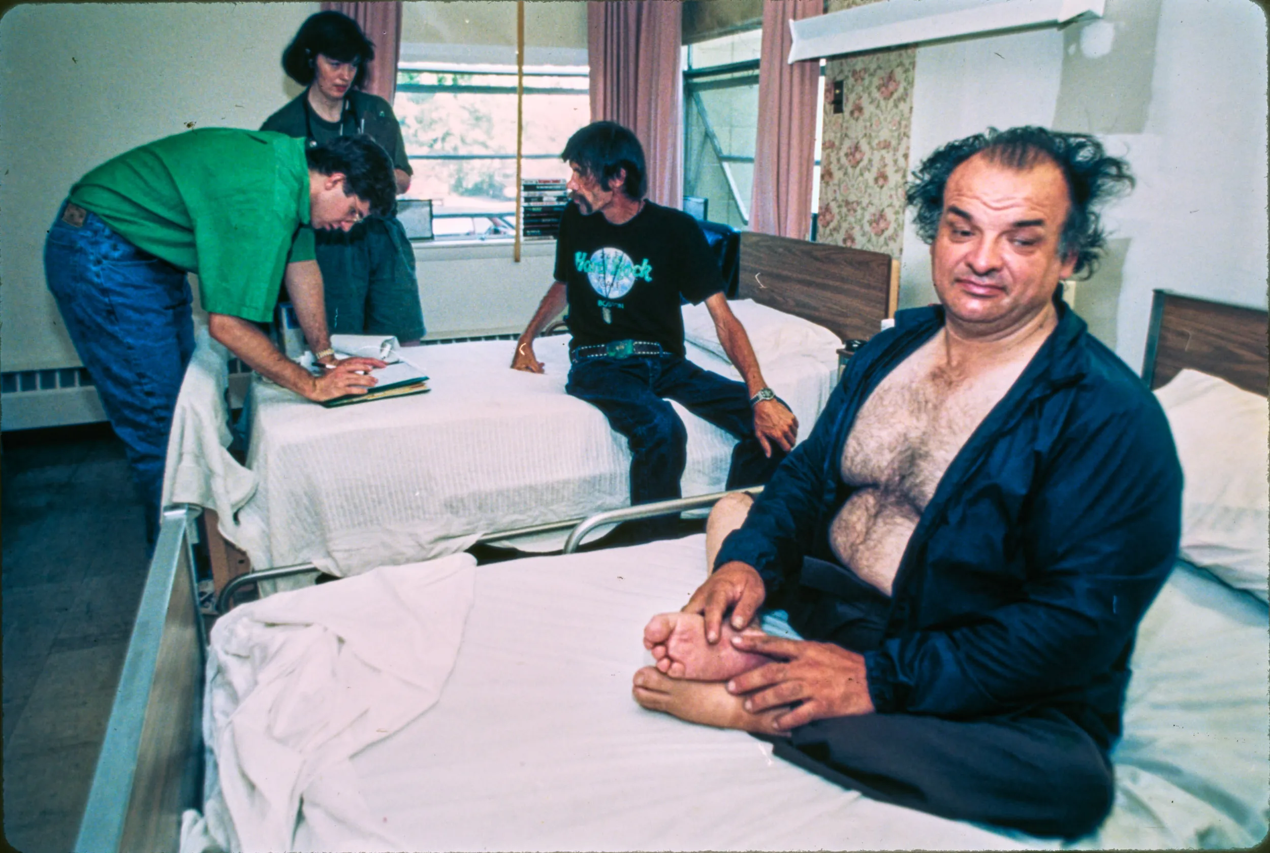 Homeless End Care — Dr. Martin Donohoe, left, and Dr. Robin Cuddy speak to homeless patient William Oakes, sitting on his bed in background, who is living with end stage AIDS at the Barbara M. McInnis Home in Boston on July 12, 1993. “Boston Health Care For The Homeless” patient Bill Latham, seated on his bed foreground, has been struggling with Polio since childhood and was just seen by doctors Donohoe and Cuddy. In addition to full-time doctor and 24-hour nursing care, respite guests have access to social services workers, physical and occupational therapists and drug and alcohol treatment programs.  (AP Photo/Stephan Savoia)  © The Associated Press